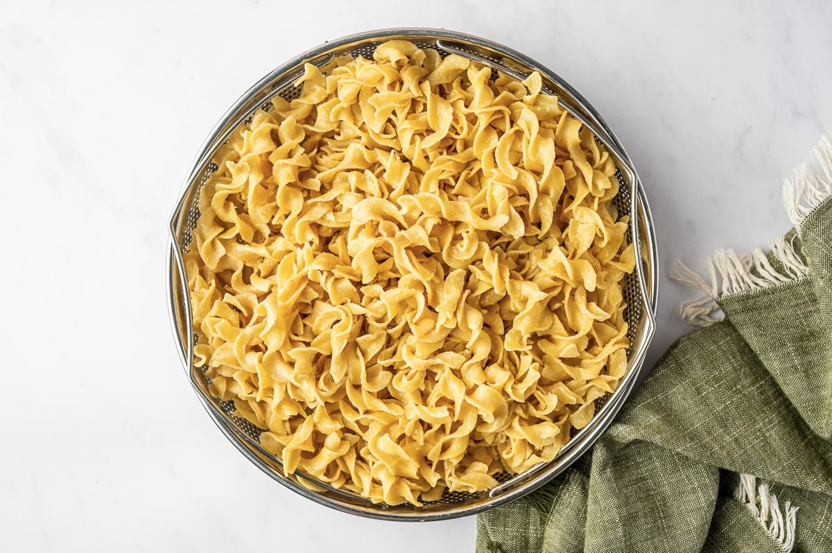 Cooked wide egg noodles draining in a large metal colander after being boiled separately for homemade chicken noodle soup.