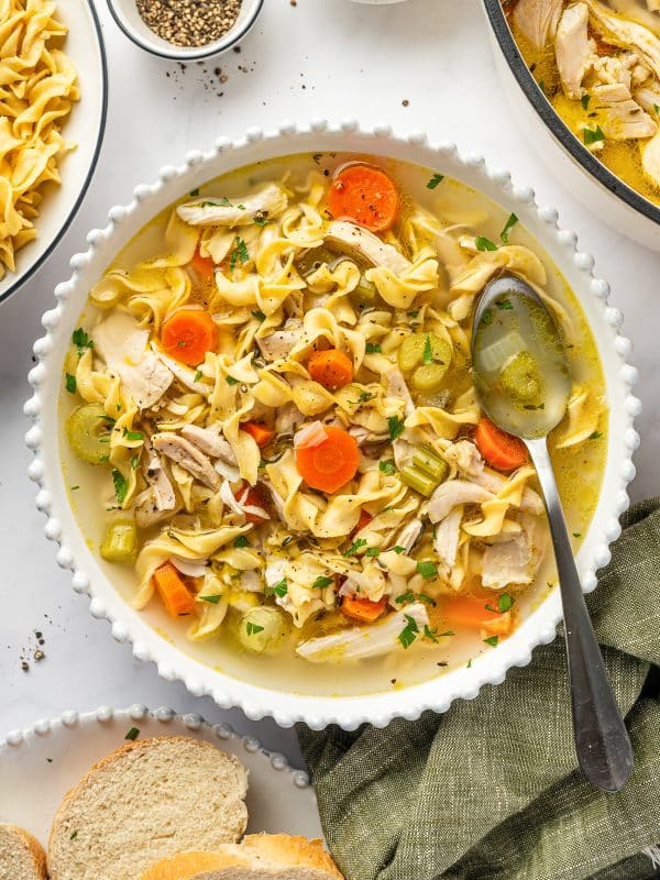 A bowl of homemade chicken noodle soup filled with wide egg noodles, shredded chicken, sliced carrots, celery, onions, herbs, and golden broth, garnished with parsley and black pepper.