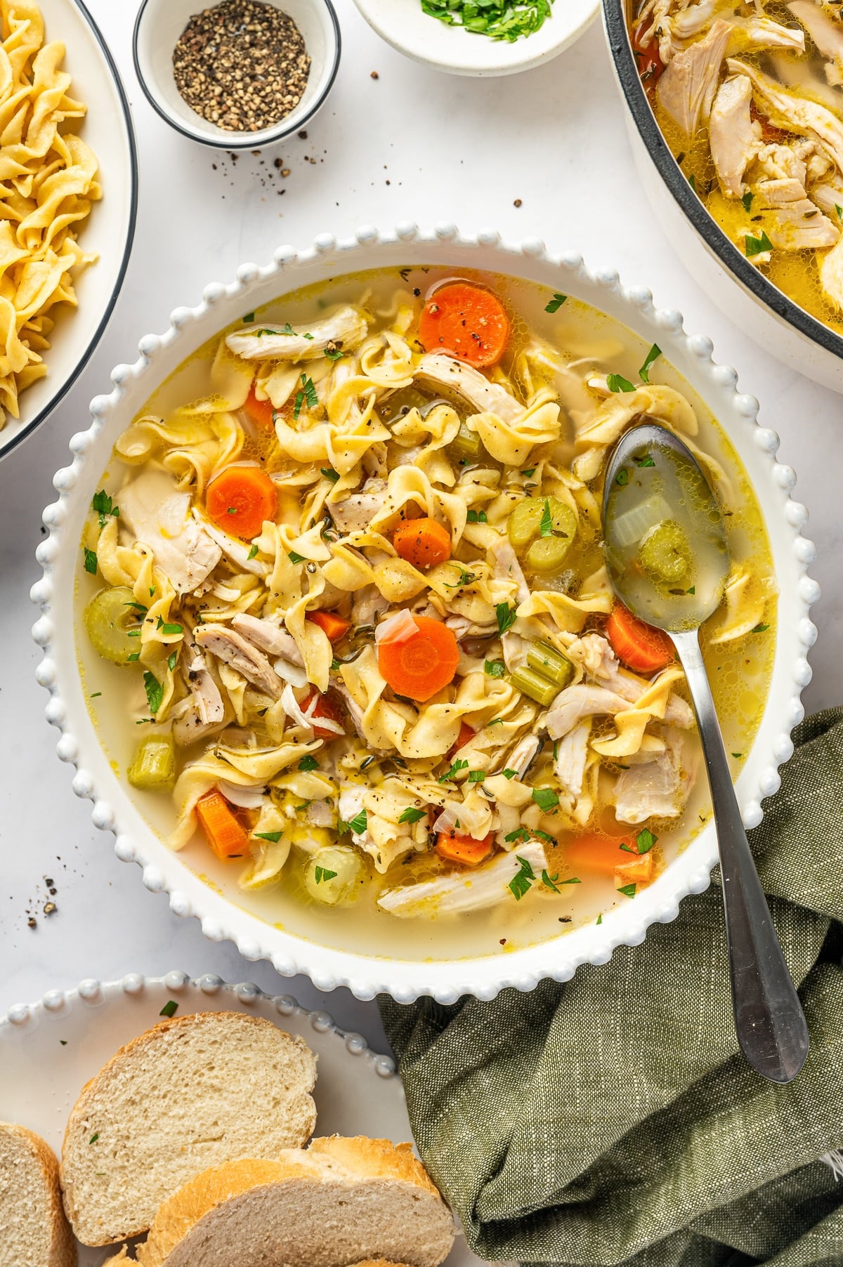 A bowl of homemade chicken noodle soup filled with wide egg noodles, shredded chicken, sliced carrots, celery, onions, herbs, and golden broth, garnished with parsley and black pepper.