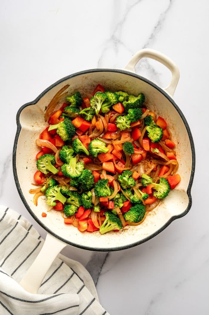 Overhead image of chopped red bell pepper, broccoli florets, and caramelized onions cooking together in a cream-colored enameled cast iron braiser on a white marble surface. The vegetables are spread across the pan, and a white kitchen towel with thin black stripes is visible underneath the lower left edge.