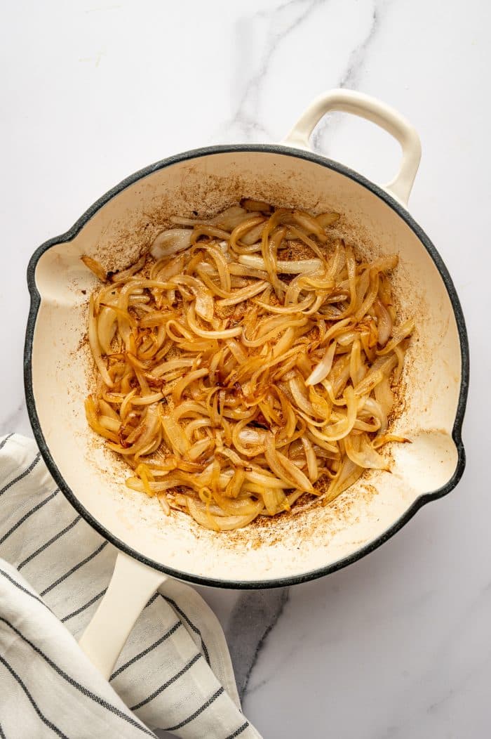 Overhead image of thinly sliced yellow onions cooking in a cream-colored enameled cast iron braiser with dual handles on a white marble surface. The onions are soft and golden brown with browned bits around the bottom and sides of the pan. A white kitchen towel with thin black stripes is tucked under the lower left side of the pan.