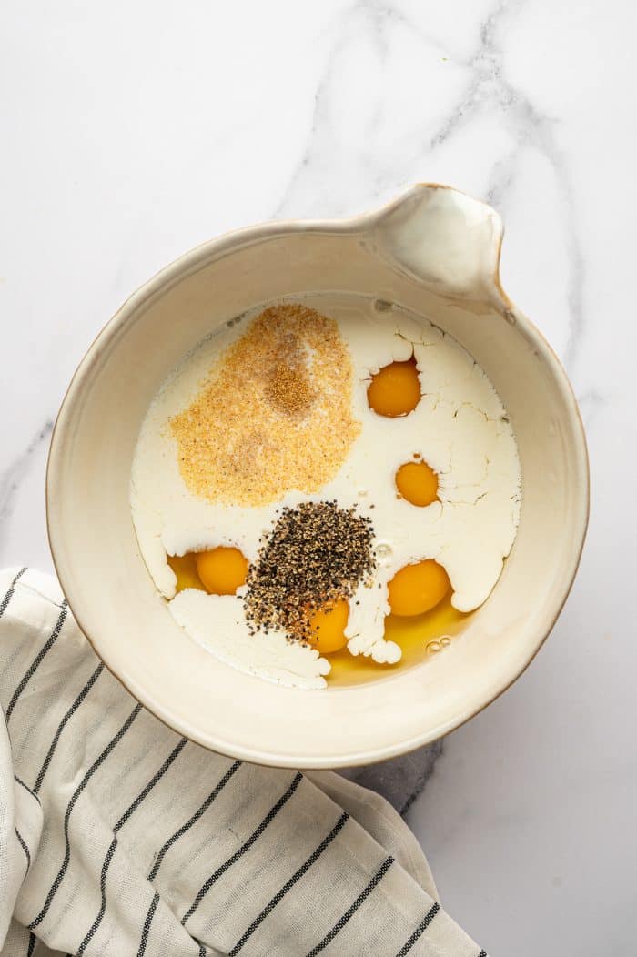 Overhead image of a cream-colored ceramic mixing bowl on a white marble surface containing cracked raw eggs, heavy cream or half-and-half, a mound of black pepper, and a sprinkle of garlic powder. A white kitchen towel with thin black stripes is placed along the lower left side of the bowl.