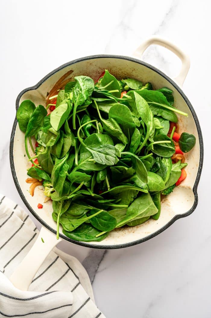 Overhead image of a cream-colored enameled cast iron braiser filled with cooked broccoli, red bell pepper, and onions, topped with a large pile of fresh baby spinach leaves. The pan sits on a white marble surface with a white kitchen towel with thin black stripes at the lower left.