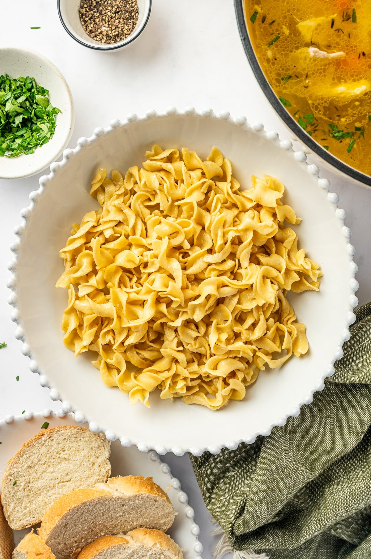 A large white serving bowl filled with cooked wide egg noodles beside a pot of chicken soup, with sliced bread, chopped parsley, and black pepper nearby.