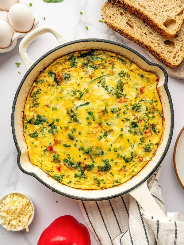 Overhead image of a baked egg frittata in a cream-colored enameled cast iron braiser on a white marble surface. The frittata is golden yellow with visible broccoli florets, spinach, red bell pepper, and melted cheese, and is garnished with chopped chives. Surrounding the pan are sliced seeded bread on a wooden board, a red bell pepper, a small white bowl of shredded cheese, a carton of white eggs, and a few scattered chive pieces and spinach leaves. A white kitchen towel with thin black stripes sits under the pan.