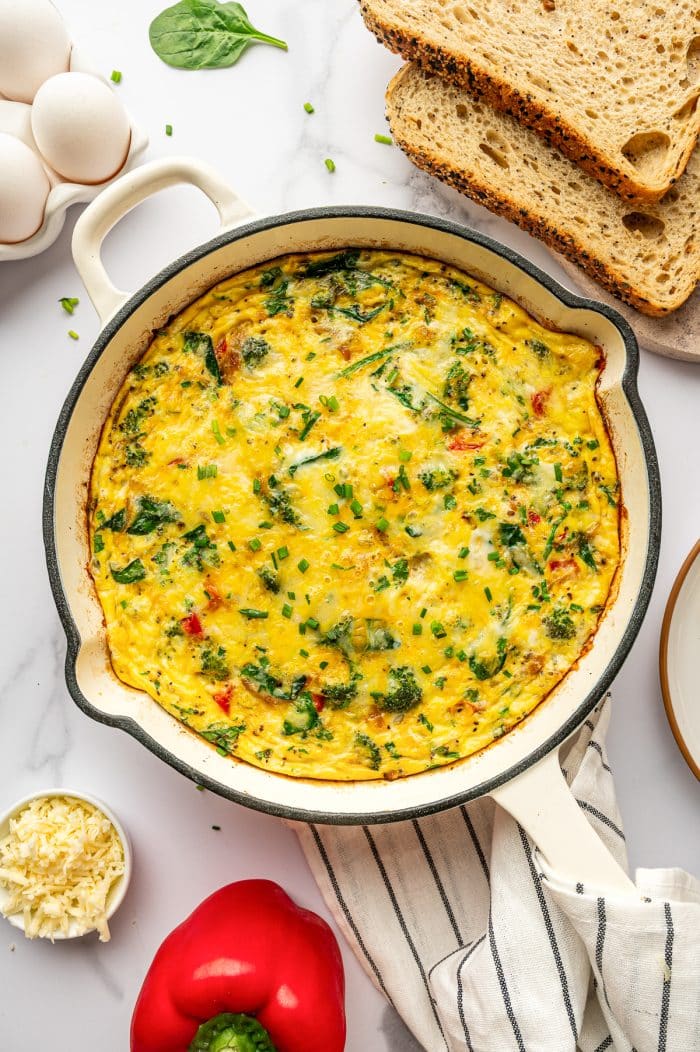 Overhead image of a baked egg frittata in a cream-colored enameled cast iron braiser on a white marble surface. The frittata is golden yellow with visible broccoli florets, spinach, red bell pepper, and melted cheese, and is garnished with chopped chives. Surrounding the pan are sliced seeded bread on a wooden board, a red bell pepper, a small white bowl of shredded cheese, a carton of white eggs, and a few scattered chive pieces and spinach leaves. A white kitchen towel with thin black stripes sits under the pan.