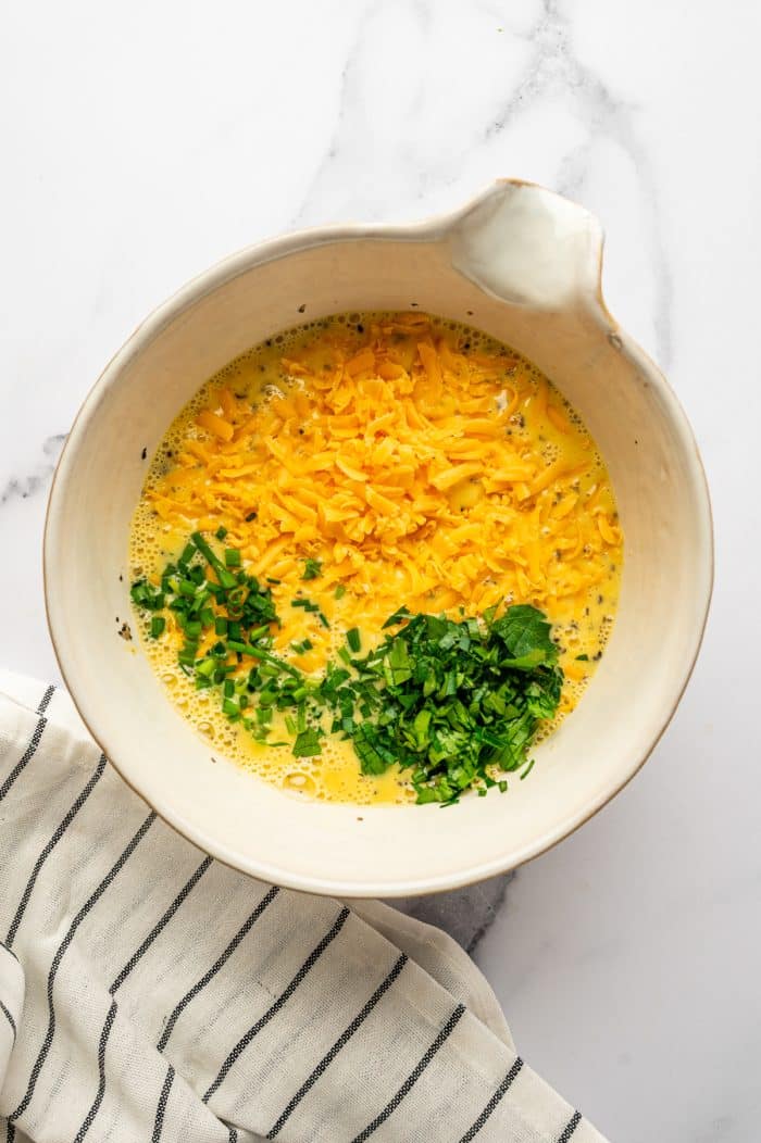 Overhead image of a cream-colored ceramic mixing bowl on a white marble surface filled with whisked eggs and cream, topped with shredded sharp cheddar cheese, chopped chives, and chopped parsley. A white kitchen towel with thin black stripes sits at the lower left side of the bowl.