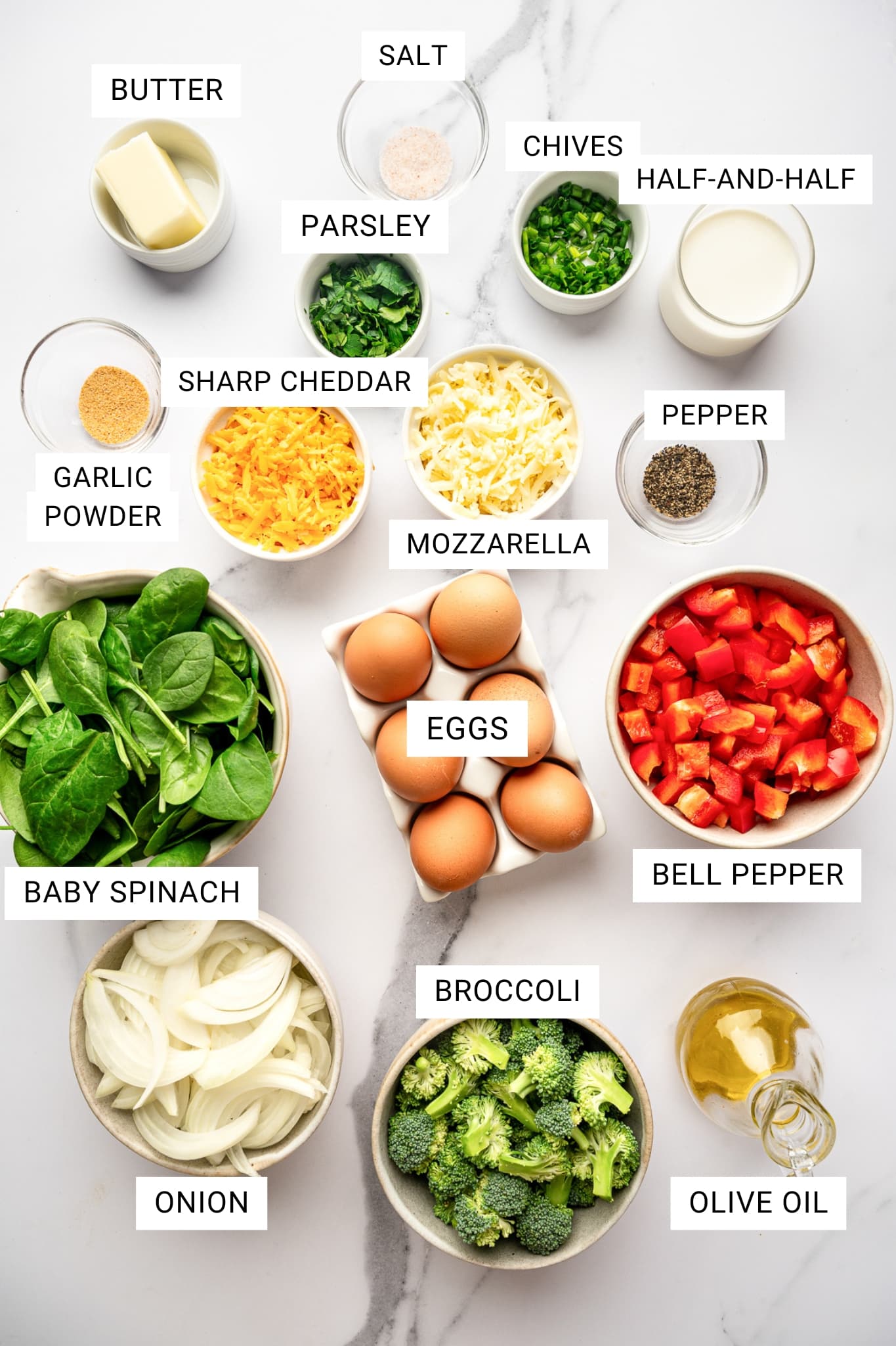 Overhead image of the ingredients for egg frittata arranged in small bowls and dishes on a white marble surface with white text labels in rectangular boxes identifying each ingredient. The labeled ingredients include butter in a white ramekin, salt in a small glass bowl, chopped chives in a white ramekin, half-and-half in a glass, chopped parsley in a white ramekin, sharp cheddar in a white bowl, mozzarella in a white bowl, black pepper in a small glass bowl, garlic powder in a small glass bowl, baby spinach in a beige bowl, eggs in a white ceramic egg holder, chopped red bell pepper in a beige bowl, sliced onion in a beige bowl, broccoli florets in a beige bowl, and olive oil in a glass bottle. No garnish beyond the arranged ingredients.