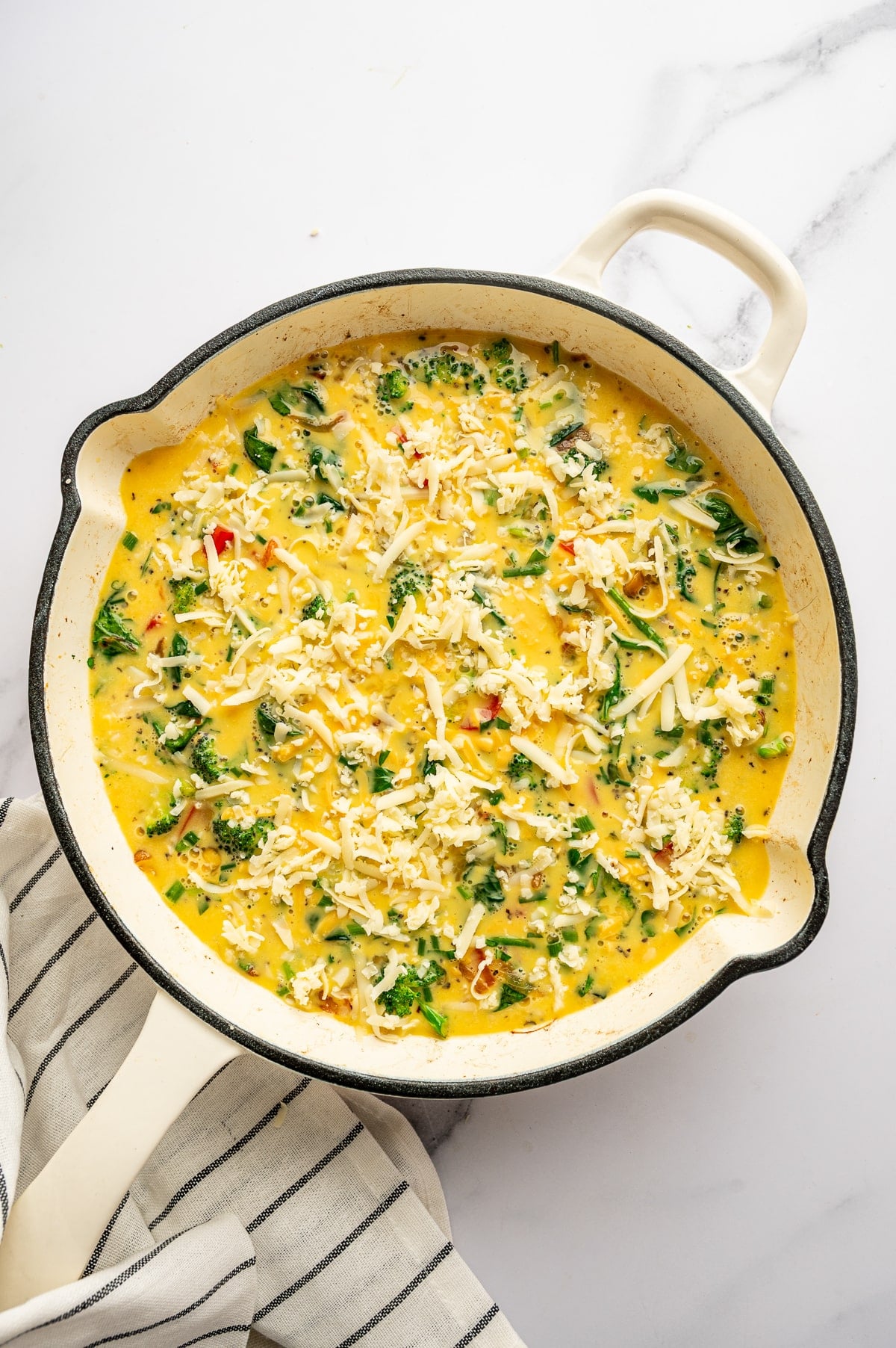Overhead image of an uncooked frittata in a cream-colored enameled cast iron braiser on a white marble surface. The pan is filled with egg mixture, broccoli, red bell pepper, wilted spinach, onions, herbs, and topped with shredded white cheese scattered across the surface. A small amount of egg mixture is visible along the pan edge, and a white kitchen towel with thin black stripes sits beneath the lower left side.