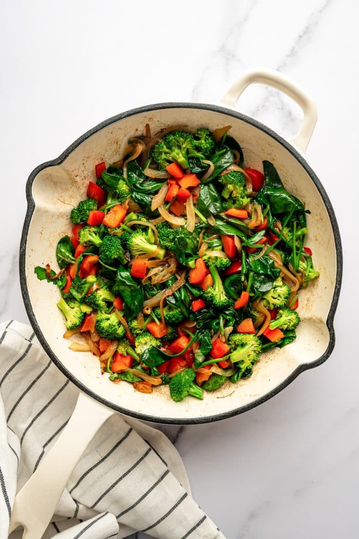 Overhead image of cooked broccoli florets, red bell pepper, caramelized onions, and wilted spinach in a cream-colored enameled cast iron braiser on a white marble surface. The vegetables are softened and mixed together, with a white kitchen towel with thin black stripes under the lower left side of the pan.