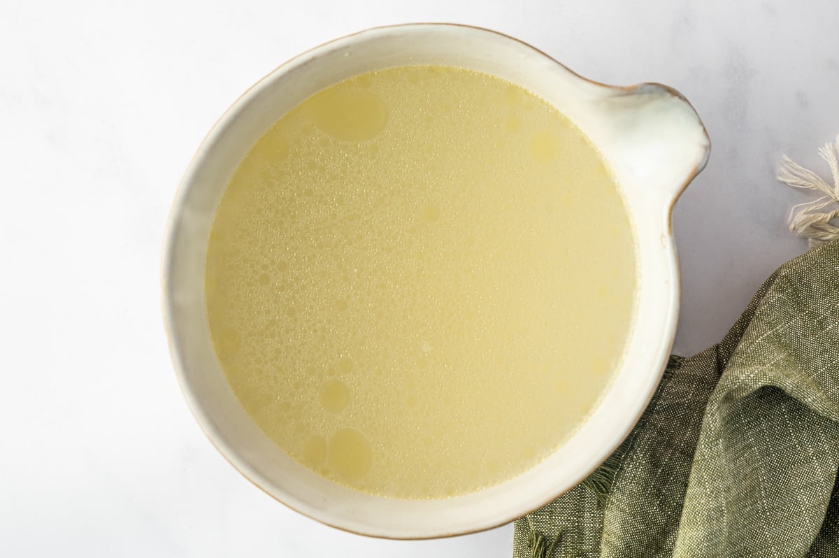 Freshly made pale golden chicken broth in a large cream-colored bowl, with small droplets of chicken fat visible across the surface.