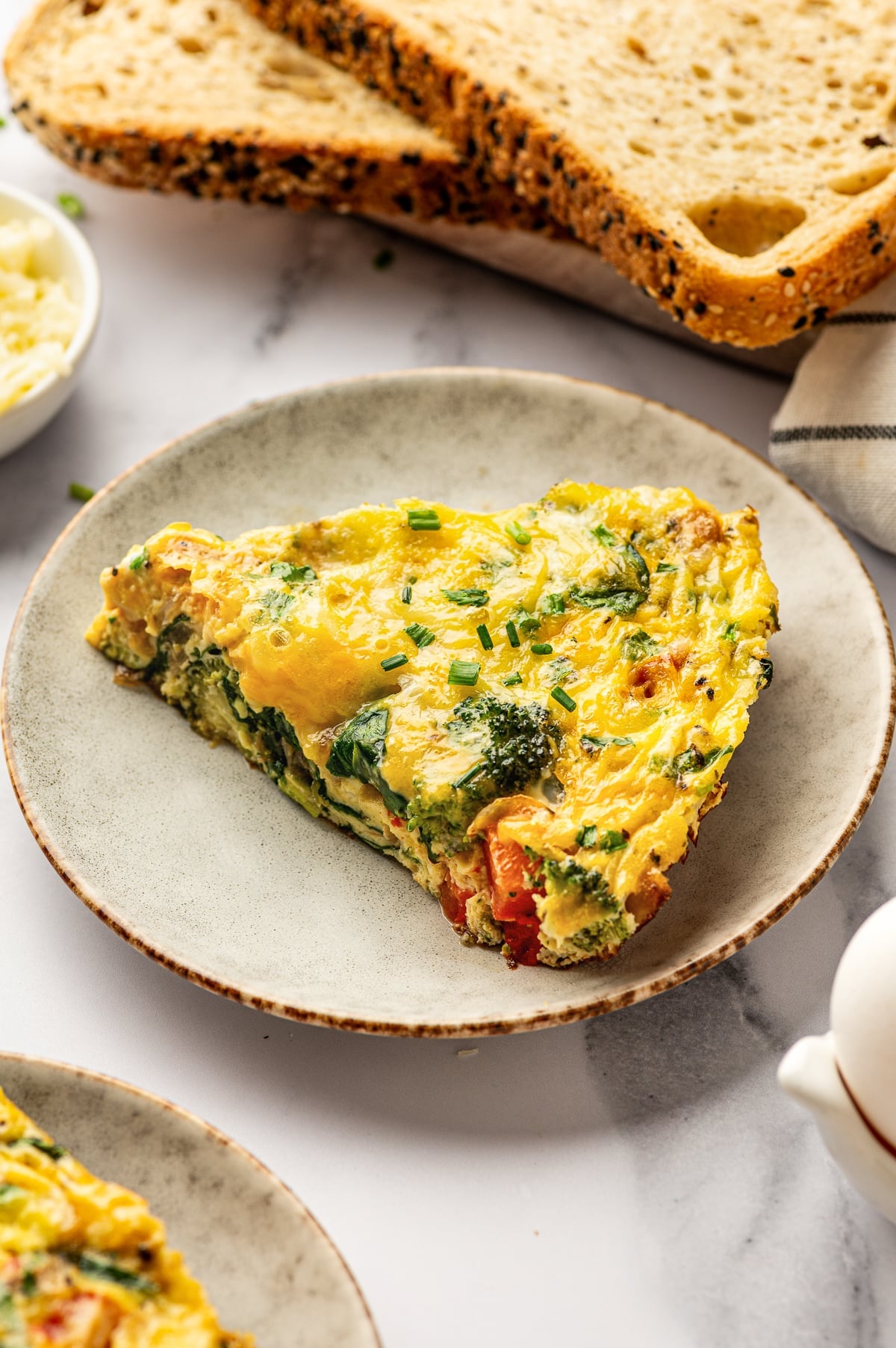Close-up image of a slice of egg frittata served on a light beige speckled ceramic plate on a white marble surface. The slice is golden yellow with melted cheese and visible broccoli, spinach, onions, and red bell pepper, topped with chopped chives. In the background are slices of seeded toast on a wooden board and a folded white kitchen towel with thin black stripes. Part of another plated slice is visible in the lower left corner, and the edge of a white ceramic egg holder appears on the right side.