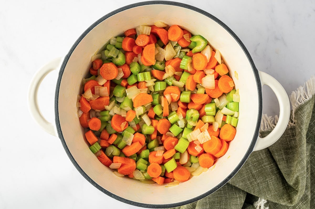 Chopped carrots, celery, and softened onions cooking together in a white Dutch oven as the vegetable base for homemade chicken noodle soup.