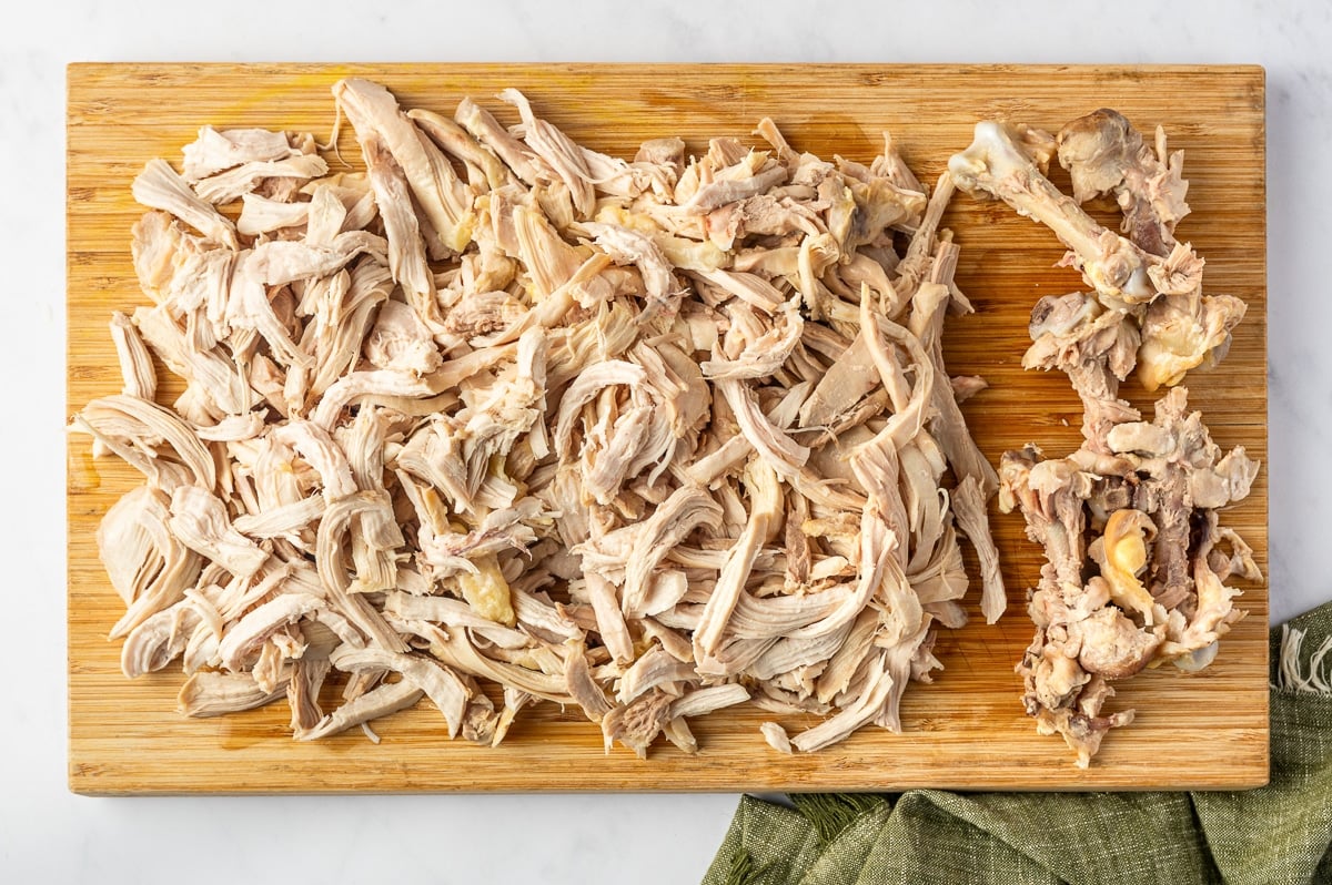 A large pile of shredded cooked chicken on a wooden cutting board with the removed chicken bones, skin, and scraps grouped separately on the right.