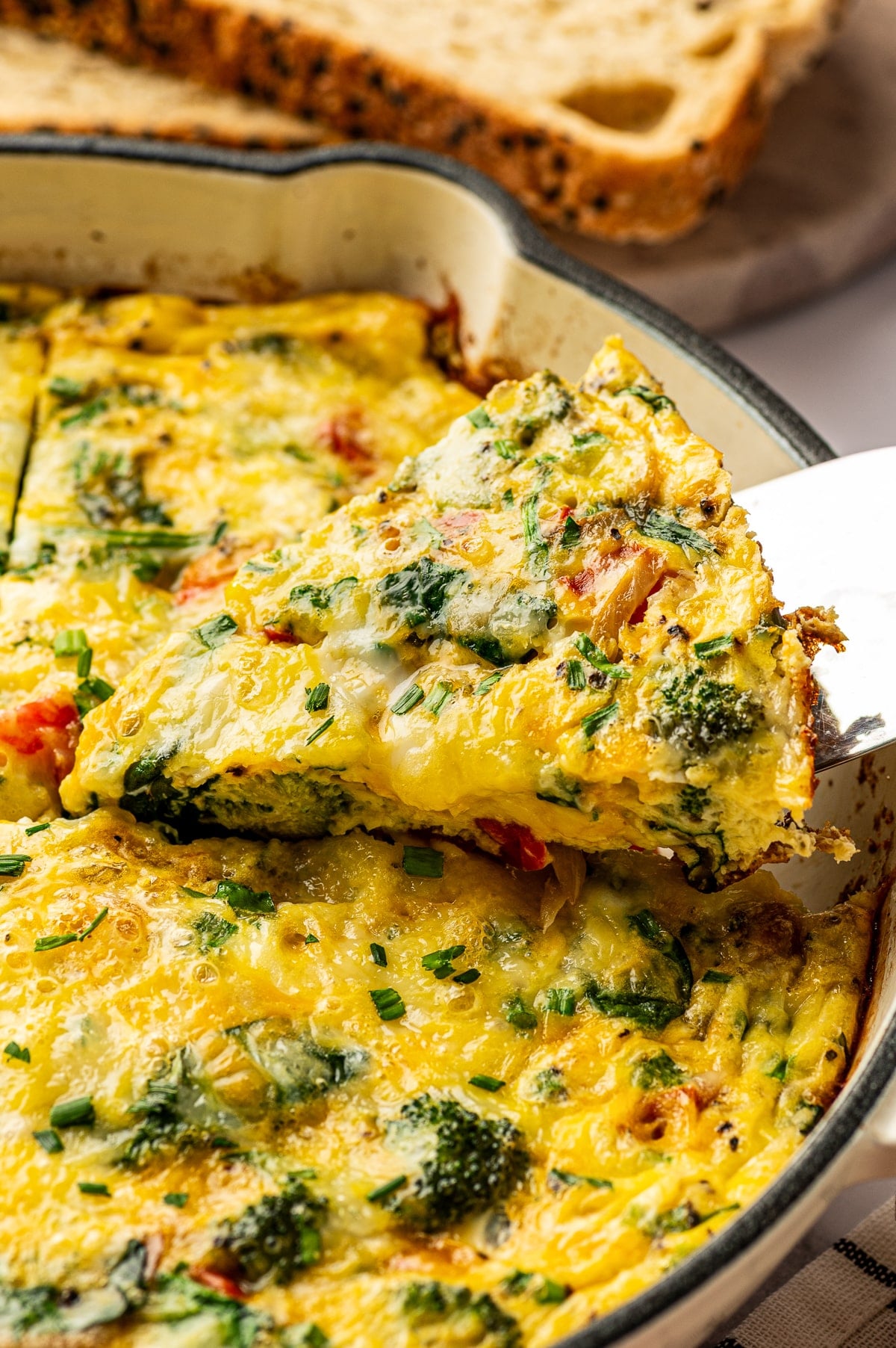 Close-up image of a thick slice of egg frittata being lifted from a cream-colored enameled cast iron braiser with a metal spatula. The frittata slice is golden yellow with melted cheese and visible broccoli, spinach, onions, and red bell pepper. More of the baked frittata remains in the pan below, topped with chopped chives. Seeded bread on a wooden board is blurred in the background.