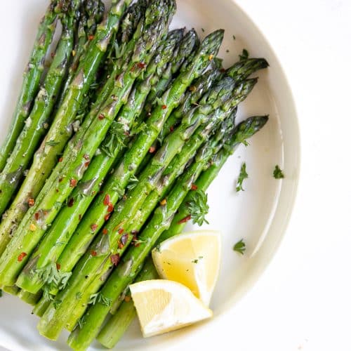 Roasted asparagus in a large flat white serving dish garnished with parsley and served with lemon.