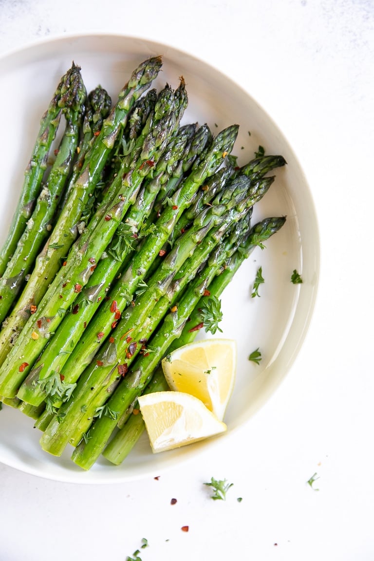 Roasted asparagus in a large flat white serving dish garnished with parsley and served with lemon.