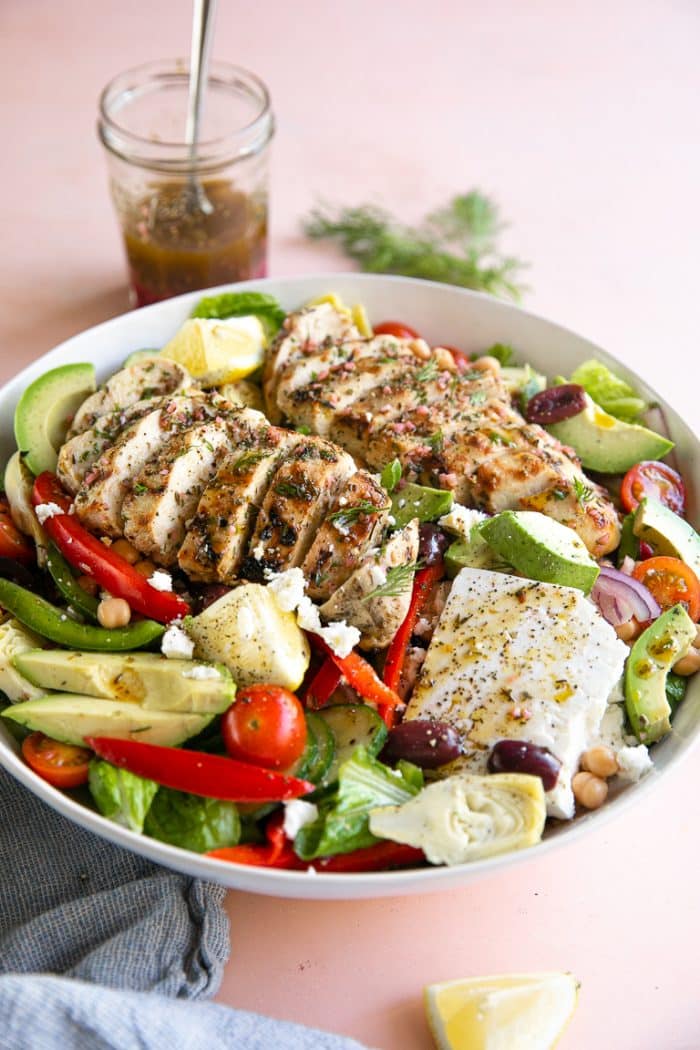 Large white serving bowl filled with lettuce, avocado, feta cheese, bell pepper, olives, and grilled Greek chicken.
