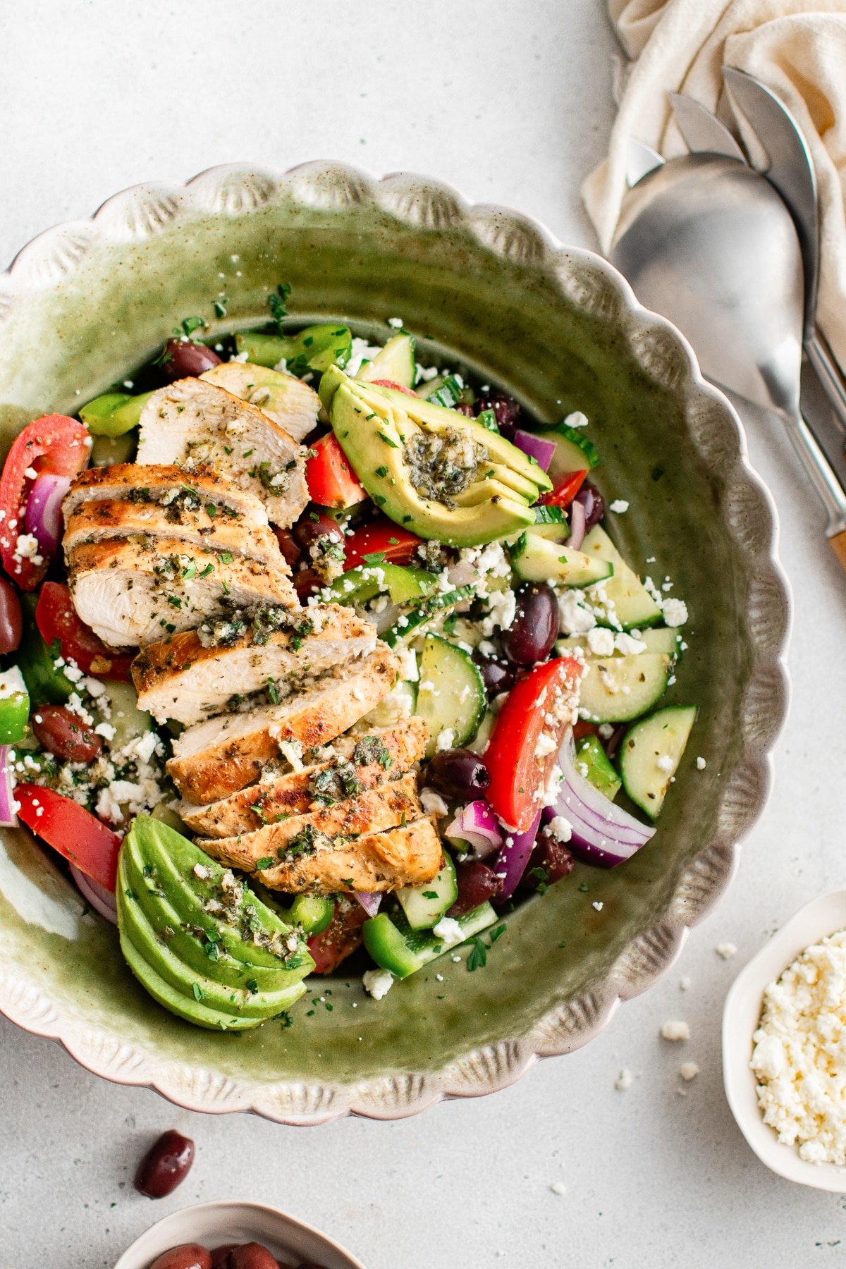 Sliced Greek chicken on a bed of Greek salad with avocado and feta in a green bowl.