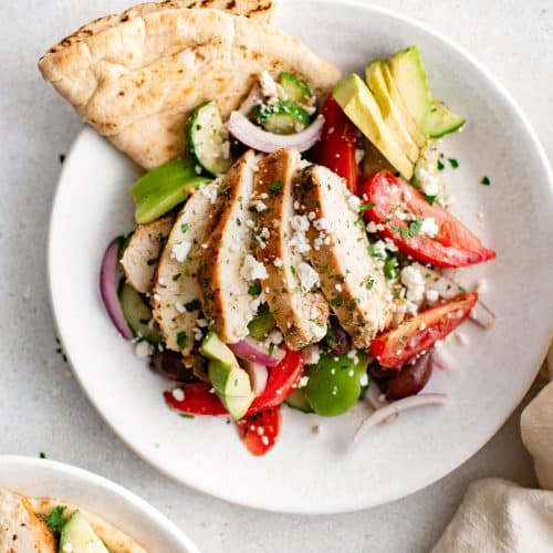 Overhead view of sliced chicken breast on Greek salad with pita and avocado.