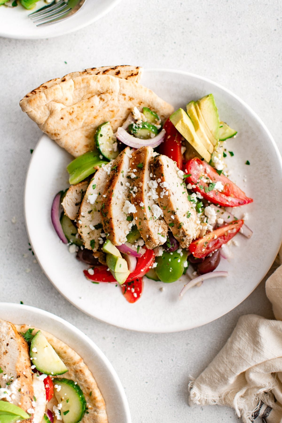 Overhead view of sliced chicken breast on Greek salad with pita and avocado.
