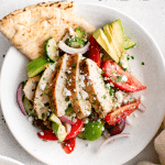 Overhead shot of sliced Greek chicken salad with avocado and pita on a white plate.