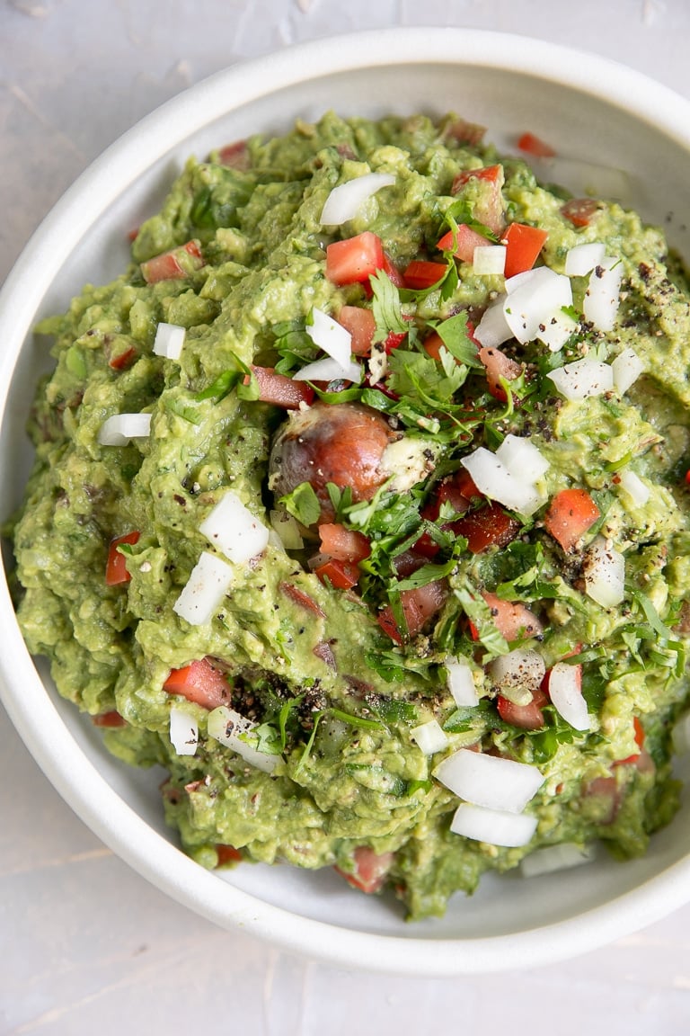 Overhead image of guacamole in a white serving bowl garnished with chopped white onion and tomato.