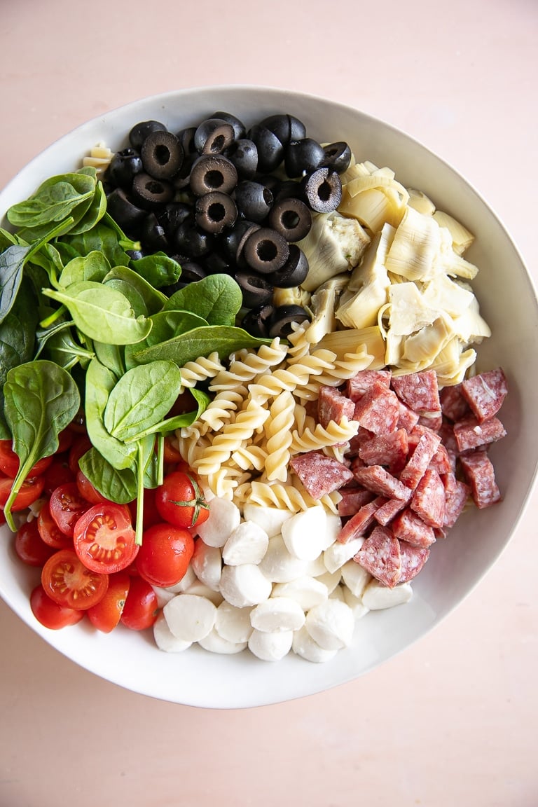 Large white bowl filled with artichokes, mozzarella, salami, cherry tomatoes, fresh spinach, olives, and pasta noodles.