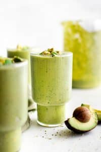 A pale green avocado smoothie in a tall clear ribbed glass, topped with diced avocado and hemp hearts, styled on a bright white surface with a halved avocado and pit in the foreground and blurred glasses of smoothie in the background.