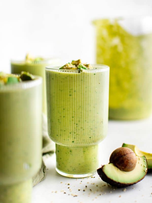 A pale green avocado smoothie in a tall clear ribbed glass, topped with diced avocado and hemp hearts, styled on a bright white surface with a halved avocado and pit in the foreground and blurred glasses of smoothie in the background.