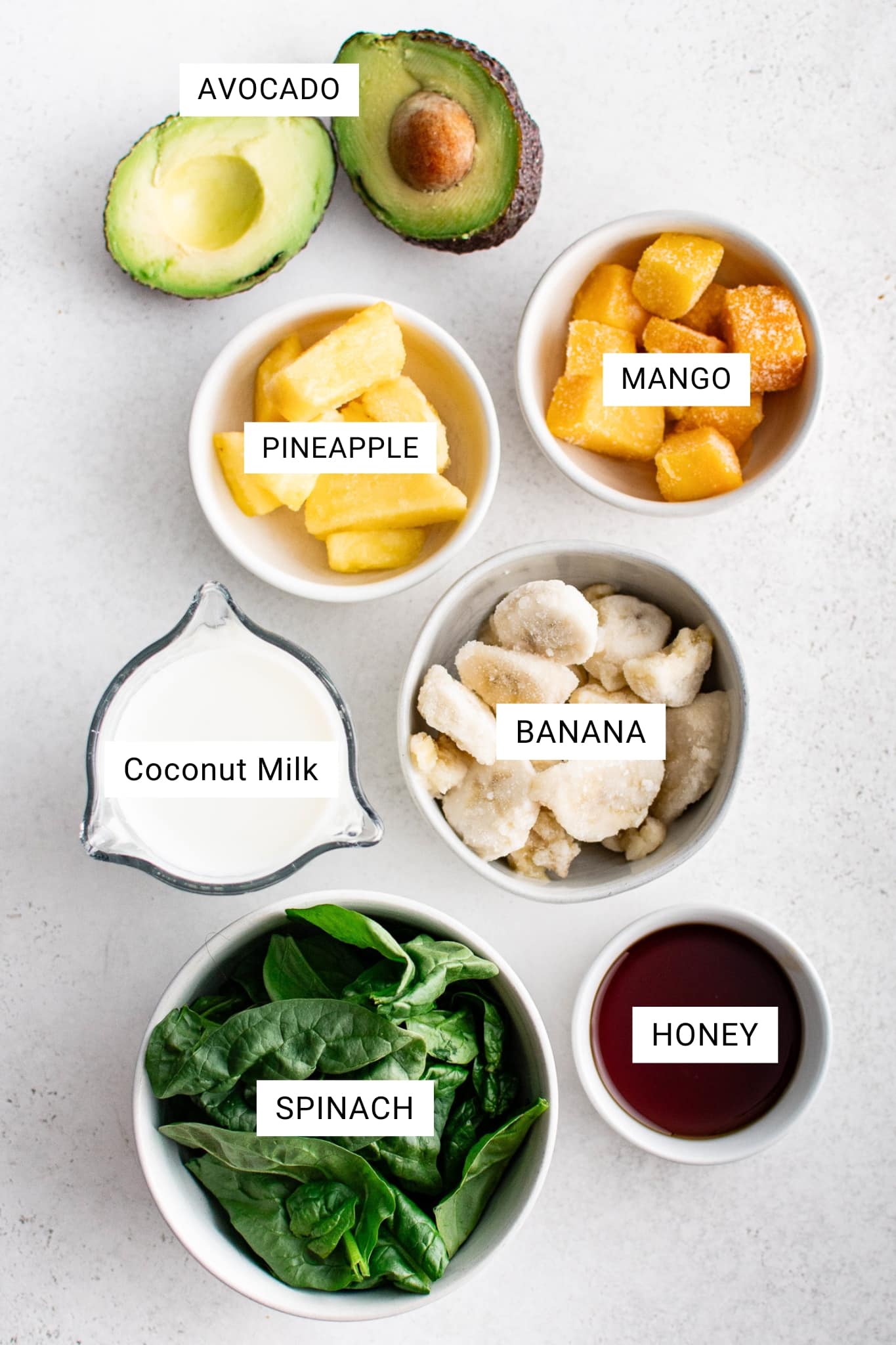 Overhead flat lay on a light gray surface showing avocado halves, frozen pineapple in a white bowl labeled “PINEAPPLE,” frozen mango in a white bowl labeled “MANGO,” frozen sliced banana in a gray bowl labeled “BANANA,” a glass measuring cup of coconut milk labeled “Coconut Milk,” fresh spinach in a white bowl labeled “SPINACH,” and honey in a small white bowl labeled “HONEY,” with an “AVOCADO” label above the avocado.