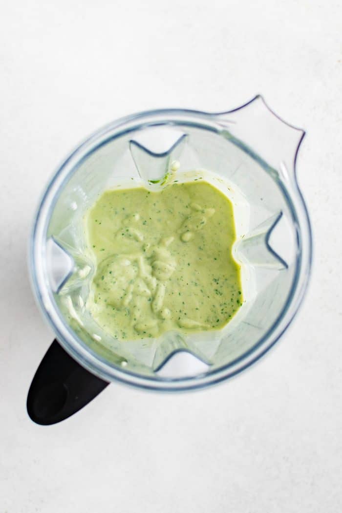 Overhead view of a clear blender jar on a light gray surface filled with pale green avocado smoothie mixture with small darker green flecks throughout.