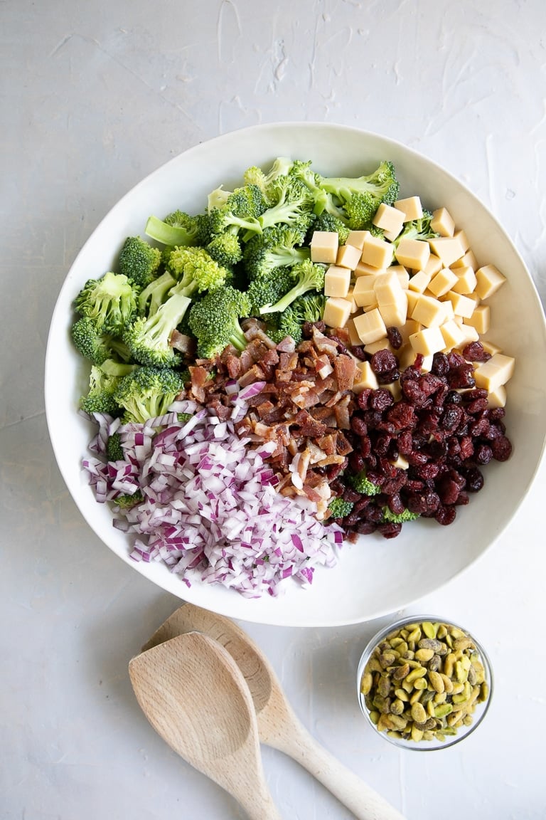 Large white mixing bowl filled with broccoli florets, dried cranberries, small cubes of smoked gouda cheese, diced red onion, and crumbled bacon.