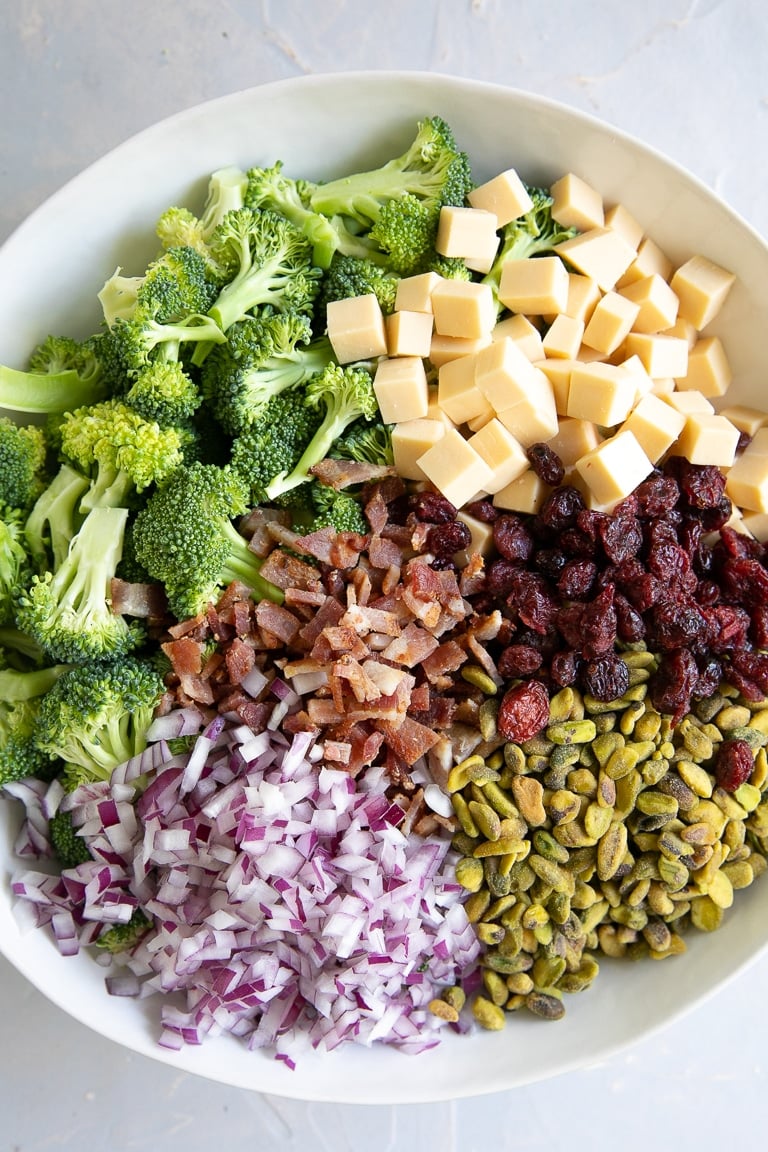 Large white mixing bowl filled with broccoli florets, dried cranberries, small cubes of smoked gouda cheese, diced red onion, crumbled bacon, and pistachios.