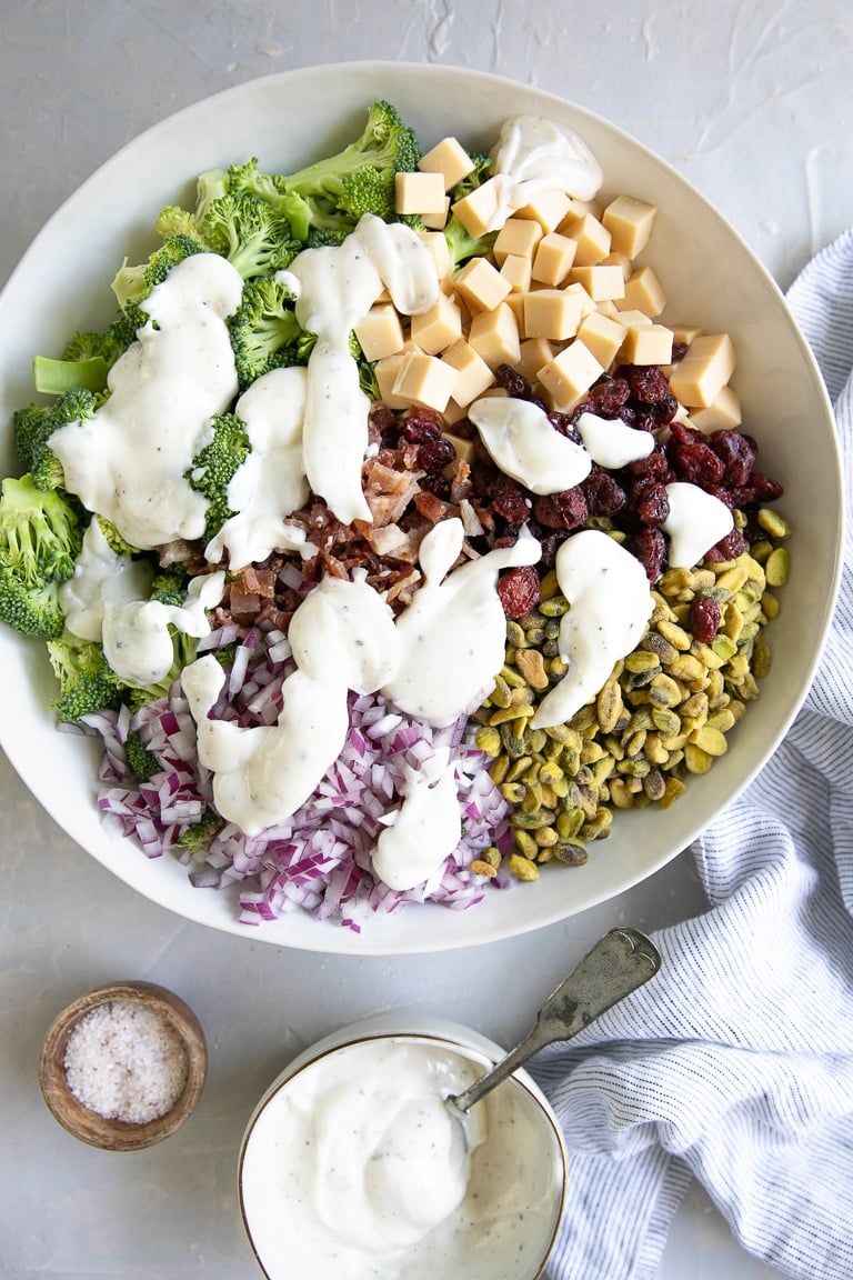 Large white mixing bowl filled with broccoli florets, dried cranberries, small cubes of smoked gouda cheese, diced red onion, crumbled bacon, pistachios, and drizzled with creamy broccoli salad dressing.