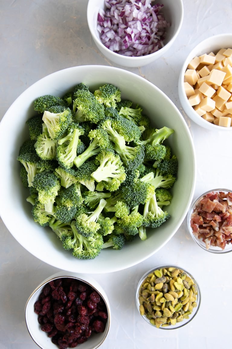 Large white bowl filled with raw broccoli florets and surrounded by five smaller bowls filled with diced red onion, cubes of smoked gouda cheese, bacon bits, pistachios, and dried cranberries.