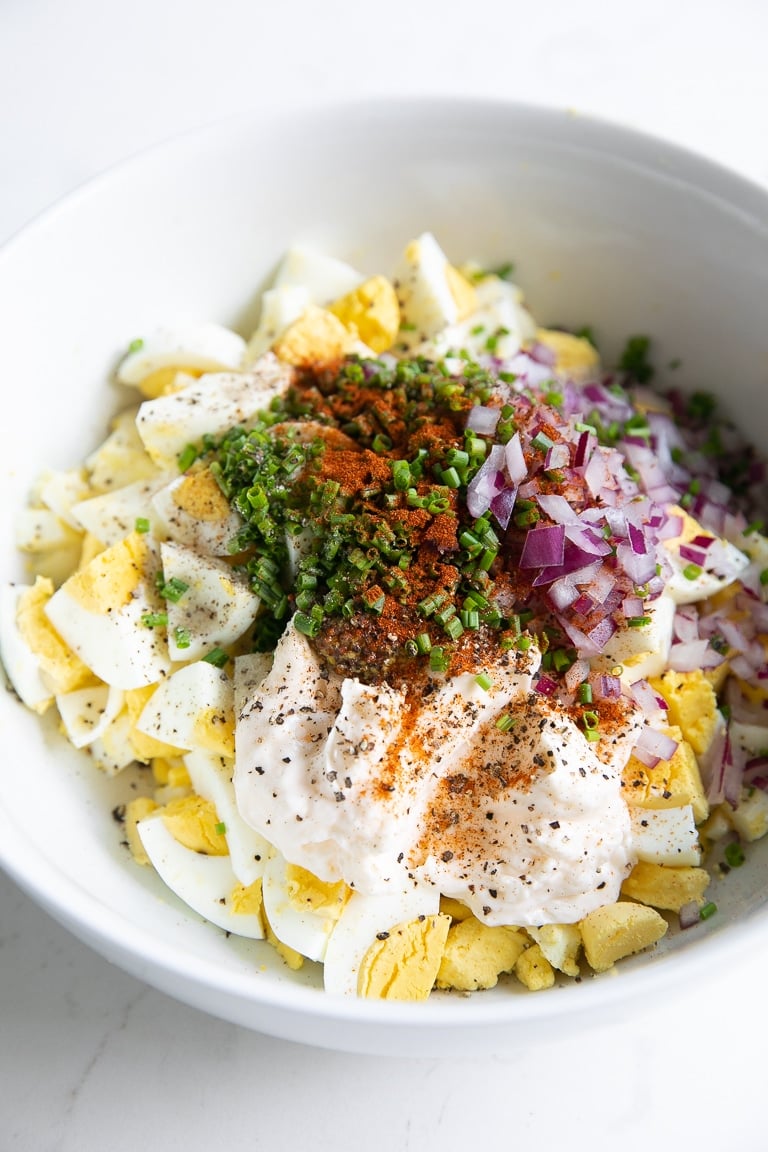 Bowl filled with hard boiled eggs, mayonnaise, parsley, diced red onion, salt, pepper, and paprika.