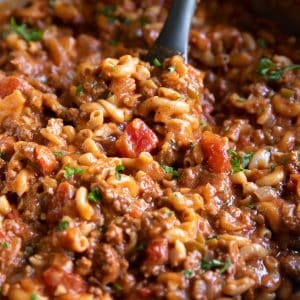 Close-up of American goulash cooking in a large stainless steel pot with elbow macaroni, ground beef, diced tomatoes, tomato sauce, and chopped parsley. A black silicone spatula with a wooden handle is lifting the pasta and beef mixture from the pot.