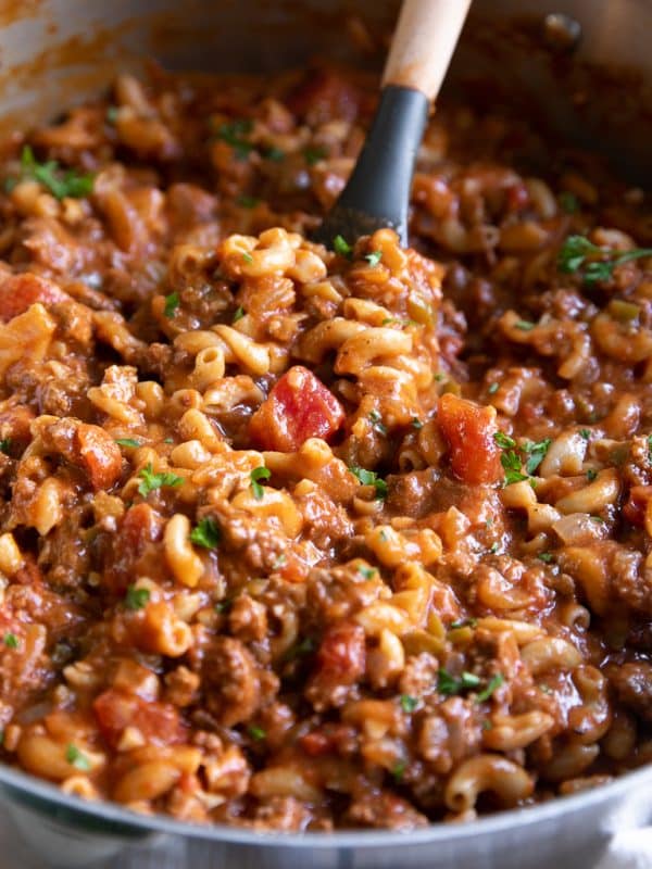 Close-up of American goulash cooking in a large stainless steel pot with elbow macaroni, ground beef, diced tomatoes, tomato sauce, and chopped parsley. A black silicone spatula with a wooden handle is lifting the pasta and beef mixture from the pot.