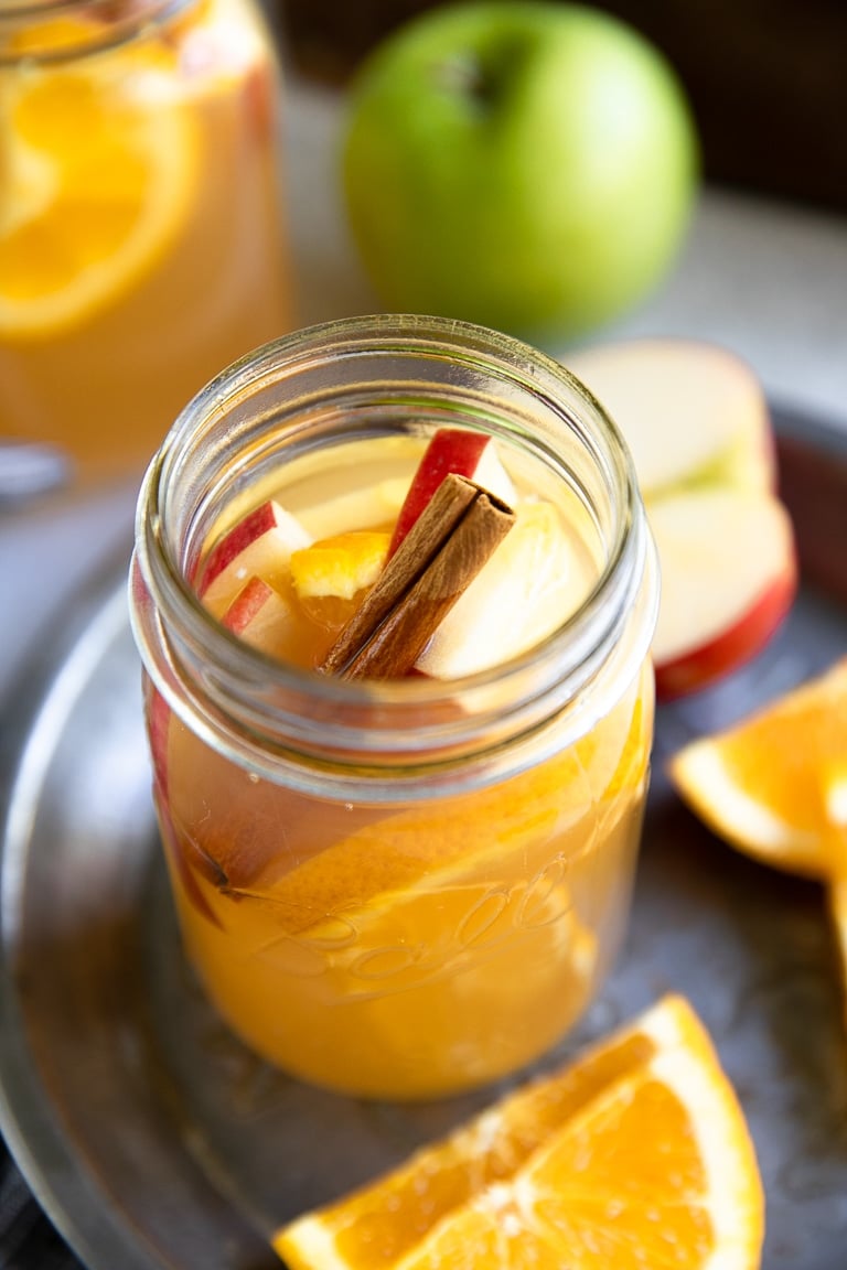 Close up image of homemade apple cider in a mason jar with apples, oranges, and a cinnamon stick.