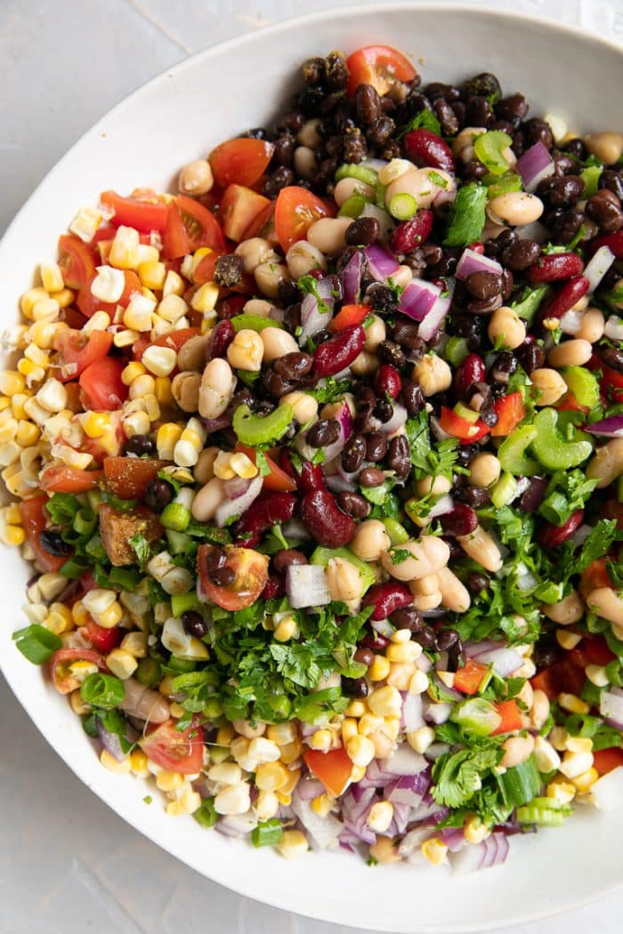 Large white mixing bowl filled with black beans, cannellini beans, kidney beans, corn, tomatoes, red onion, green onion, bell pepper, and cumin.