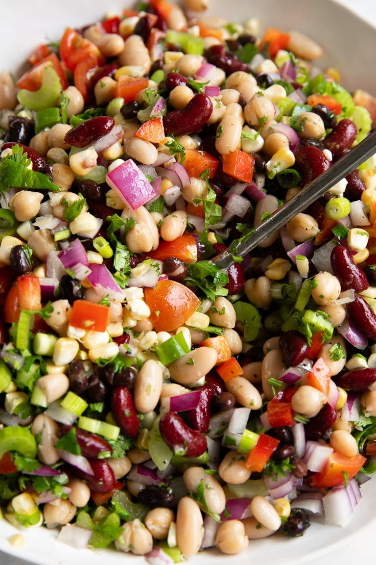 Large white mixing bowl filled with black beans, cannellini beans, kidney beans, corn, tomatoes, red onion, green onion, bell pepper, and cumin.