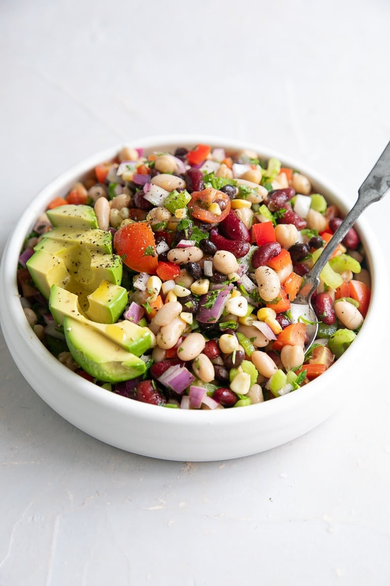 White serving bowl filled with mixed canned beans, bell pepper, cilantro, onion, tomatoes, corn, and avocado.