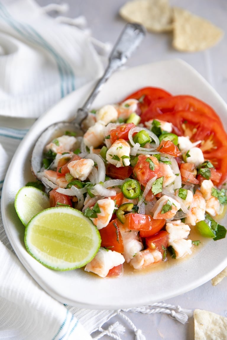 Shrimp ceviche made with onions, tomatoes, bell pepper, lime juice, and cilantro on a white serving plate with sliced tomatoes and fresh lime wedges..