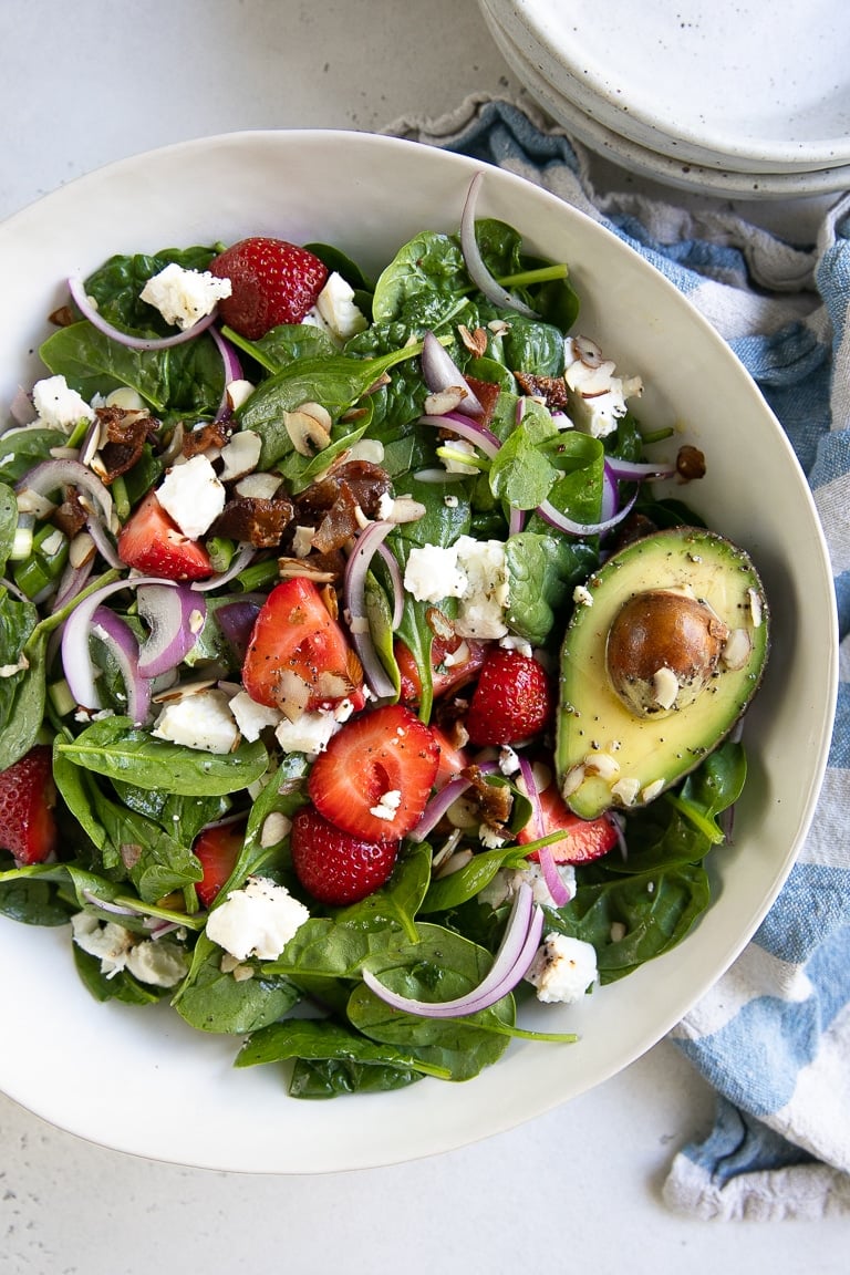 Large white salad bowl filled with tossed together spinach, strawberries, bacon, red onion, feta, green onion, slivered almonds, and green onions.