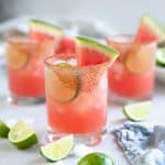 Three iced watermelon margaritas in rocks glasses garnished with fresh lime wheels and sliced watermelon.