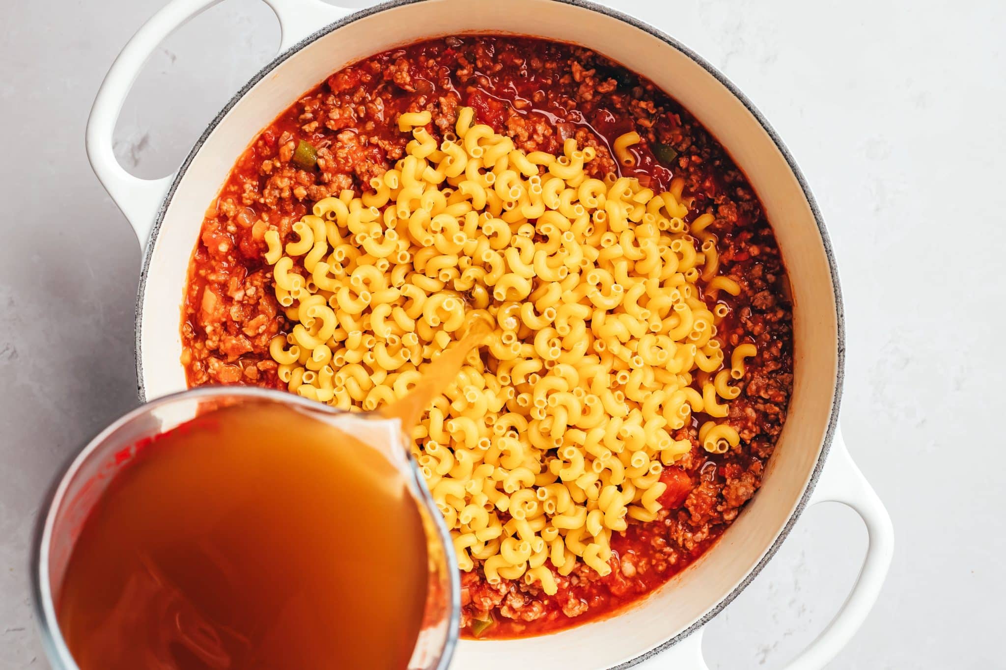 A mound of uncooked yellow elbow macaroni sits on top of a thick red tomato and ground beef sauce in a white Dutch oven. Brown beef broth is being poured from a clear glass measuring cup into the pot around the dry pasta.