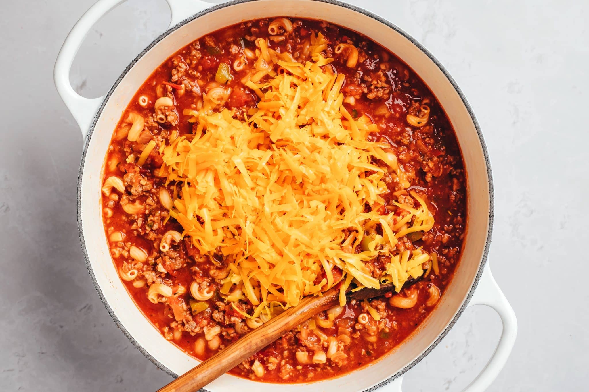 A generous pile of freshly shredded orange cheddar cheese covers the center of a Dutch oven filled with elbow macaroni, ground beef, and tomato sauce. A wooden spoon rests beneath the shredded cheese, with the red sauce and curved pasta visible around the edges.