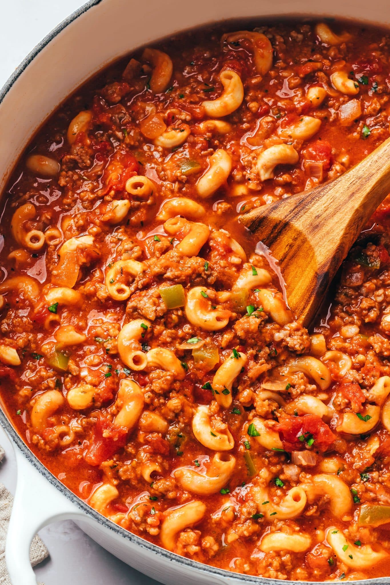 Tender elbow macaroni coated in a glossy red tomato sauce with small crumbles of ground beef, pieces of diced tomatoes, translucent onions, and green bell peppers. Chopped fresh parsley is scattered throughout the goulash, and a wooden spoon rests in the thick, saucy mixture.