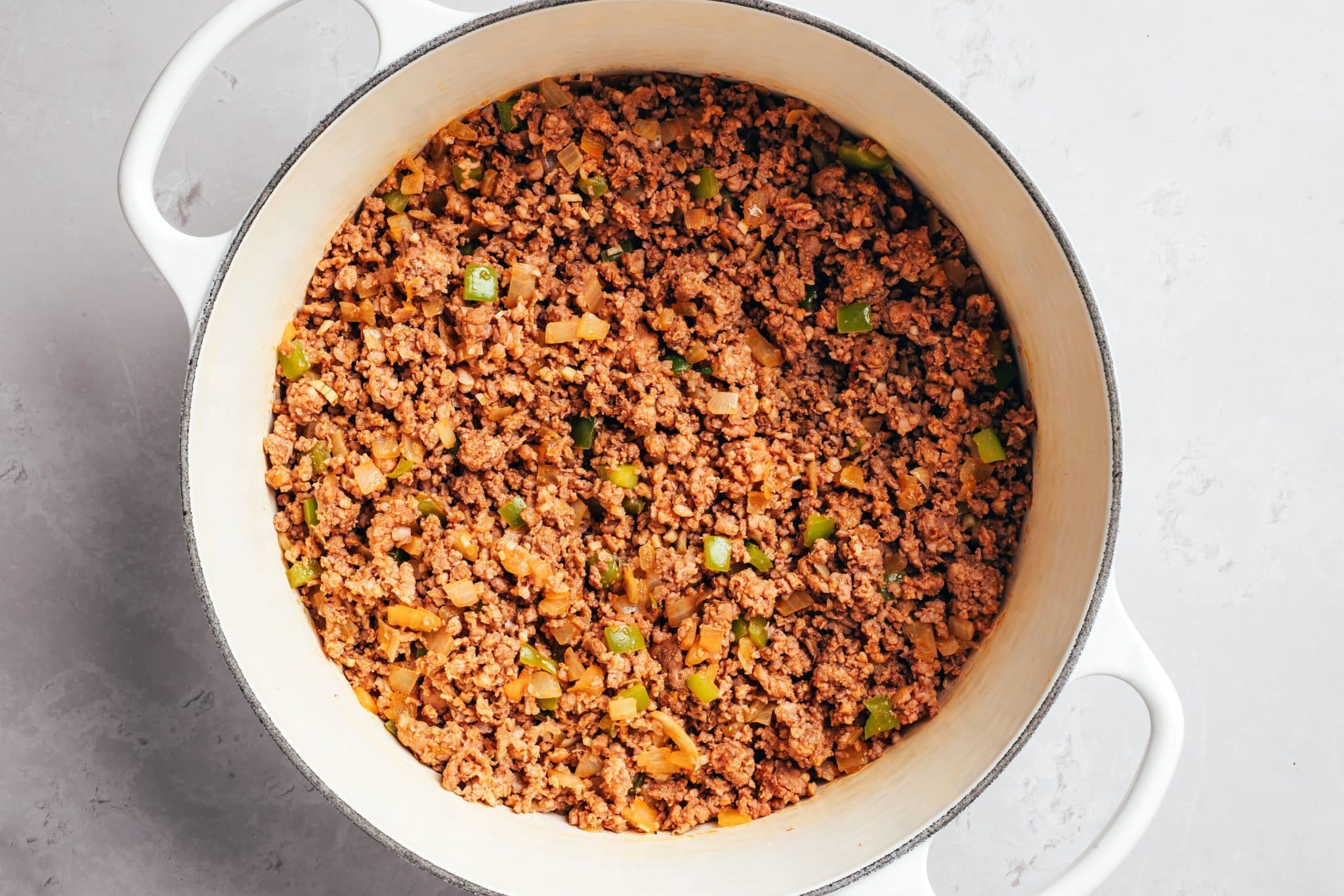 Crumbled ground beef cooked until browned and mixed with softened diced onions and green bell peppers in a white Dutch oven. The beef is broken into small, evenly sized pieces, with bits of vegetables and seasonings distributed throughout.